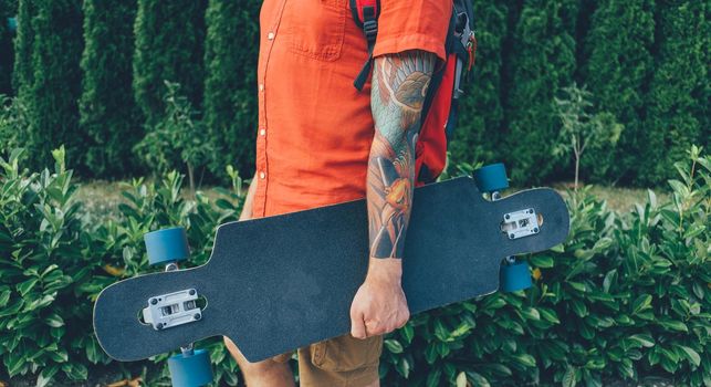 Unrecognizable tattoed young man standing with longboard in summer park.