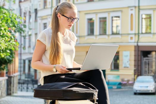 Beautiful young woman sitting outdoors using laptop, city street background. Female in business clothes, study, business, lifestyle concept