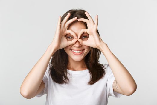 Close-up of happy smiling brunette girl looking through okay gestures, hand glasses, white background.