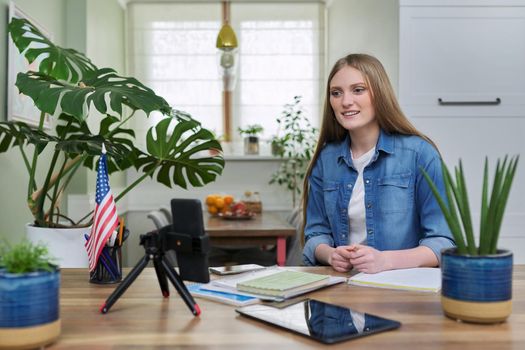 Young woman university student studying at home online remotely, e-learning. Talking female looking at smartphone screen, USA flag background, distance concept