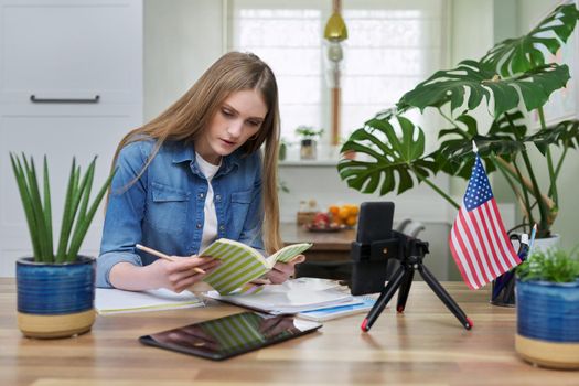 Young woman university student studying at home online remotely, e-learning. Talking female looking at smartphone screen, USA flag background, distance concept