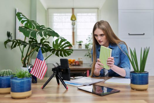 Young woman university student studying at home online remotely, e-learning. Talking female looking at smartphone screen, USA flag background, distance concept