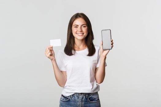 Attractive smiling girl looking satisfied and showing credit card, mobile phone screen.