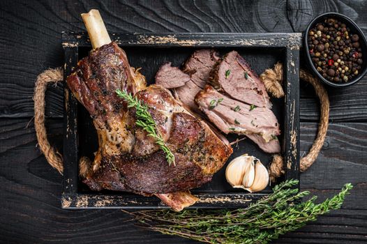 Roasted mutton lamb leg sliced in a wooden tray with meat cleaver. Black wooden background. Top view.