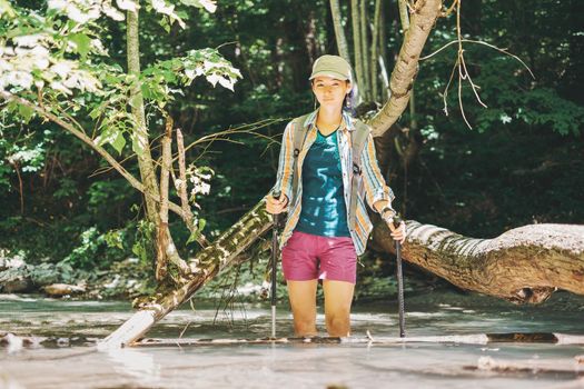 Traveler young woman with trekking poles crossing the river in summer, looking at camera.