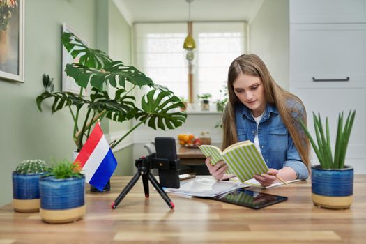 Young female university student sitting at home studying online, looking at smartphone webcam. On table flag of Netherlands, education in Holland, e-education, e-learning, technology, knowledge