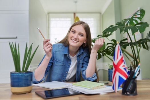 UK learning English online. Female student remotely looking at webcam taking private lessons sitting at home in kitchen, England flag background