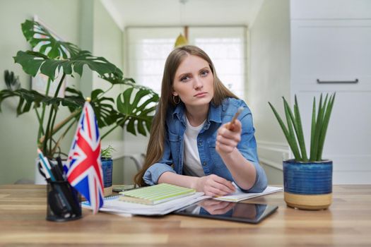 UK learning English online. Female student remotely looking at webcam taking private lessons sitting at home in kitchen, England flag background