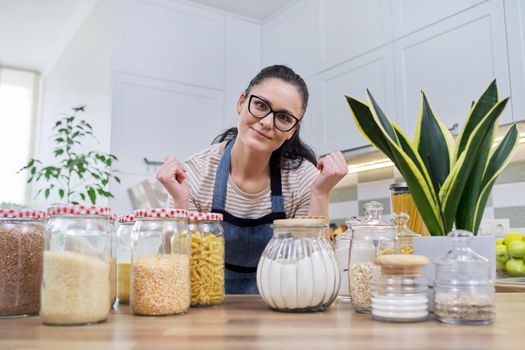 Storing food in kitchen, woman housewife in an apron with jars and containers of cereals, food, pasta, talking and looking at camera. Female blogging about organizing space in kitchen