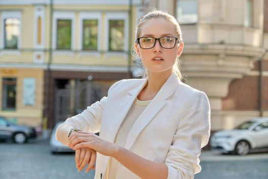 Young successful business woman looking at her wristwatch, beautiful female in glasses in white jacket on background of sunny city street