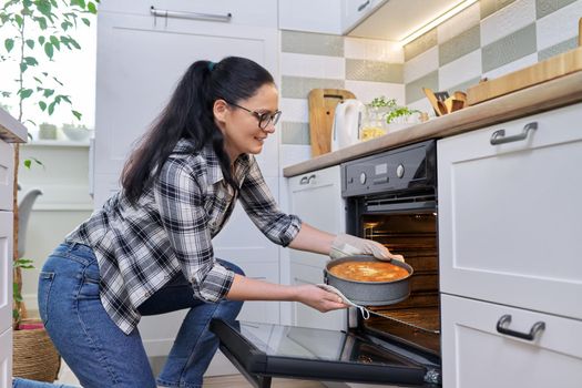 Woman at home in kitchen taking out hot apple pie from oven. Cooking at home, homemade baking, eating at home, lifestyle, hobbies and leisure