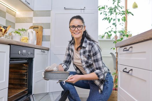 Woman at home in kitchen taking out hot apple pie from oven. Cooking at home, homemade baking, eating at home, lifestyle, hobbies and leisure