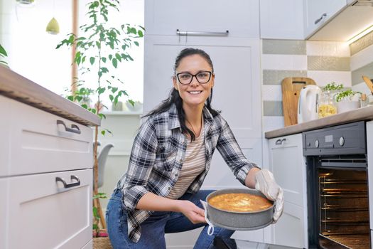 Woman at home in kitchen taking out hot apple pie from oven. Cooking at home, homemade baking, eating at home, lifestyle, hobbies and leisure