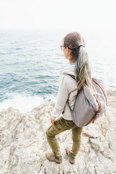 Backpacker girl standing on rocky coastline and enjoying view of sea.