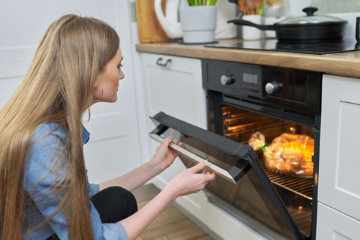 Cooking at home, roasting meat in oven, young woman putting marinated chicken in baking bag in oven, kitchen interior background