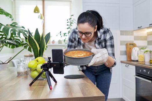 Food blog, woman blogger cooking apple pie at home, recording recipe on smartphone. Positive woman at home in kitchen talking online with friends family, using video call