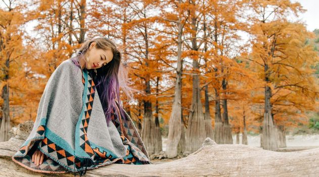 Beautiful young woman wearing in poncho resting on tree trunk on background of cypresses in autumn.