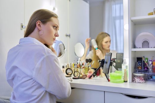 Beauty blog, young woman sitting at make-up table with mirror doing makeup hairstyle, styling her hair with brush, blonde female beauty blogger recording blog vlog on smartphone, online master class