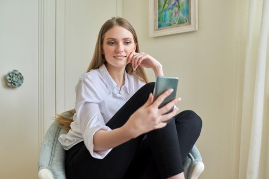 Young beautiful blonde sitting at home in chair, smiling talking using video communication on smartphone. Leisure, woman relaxing looking at phone screen