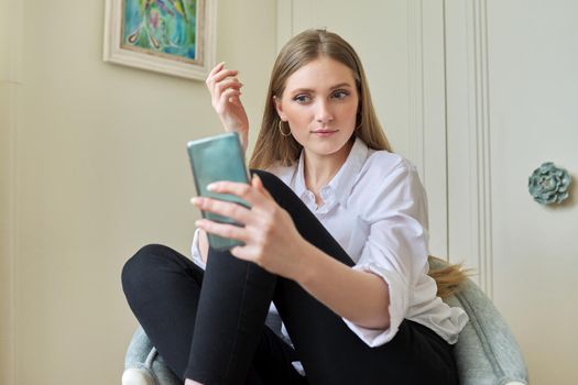 Young beautiful blonde sitting at home in chair, smiling talking using video communication on smartphone. Leisure, woman relaxing looking at phone screen