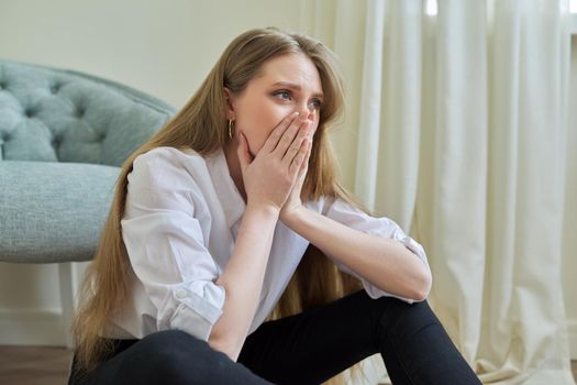 Upset crying young woman sitting at home on the floor. Negative emotions, stress, mental health, frustration, pain, people concept