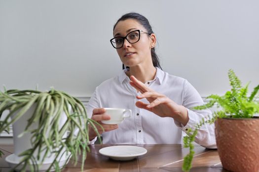 Talking positive business woman looking at camera. Middle-aged female wearing white shirt glasses sitting at table with cup of coffee, gesturing making video consultation, recording video vlog