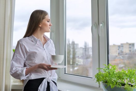 Young beautiful woman with cup of tea at home near window, blonde with long hair in white shirt looking out window, spring winter season, copy space