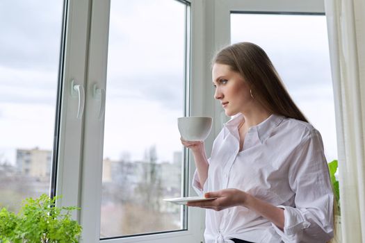 Beautiful smiling young woman with natural blond long hair holding cup coffee near panopam window. Attractive female in white shirt, winter spring season in window, copy space