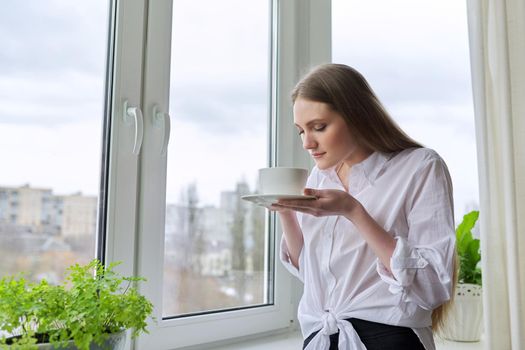 Young beautiful woman with cup of tea at home near window, blonde with long hair in white shirt looking out window, spring winter season, copy space