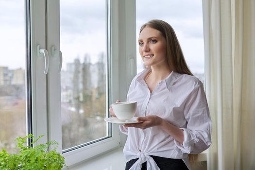 Beautiful smiling young woman with natural blond long hair holding cup coffee near panopam window. Attractive female in white shirt, winter spring season in window, copy space