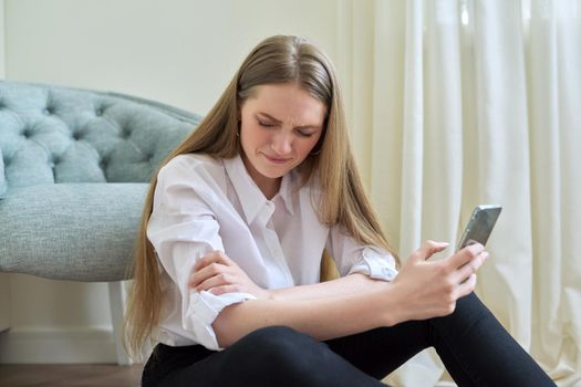 Sad crying young woman with smartphone sitting on the floor at home. Depression, bad news, problem, pandemic, illness concept