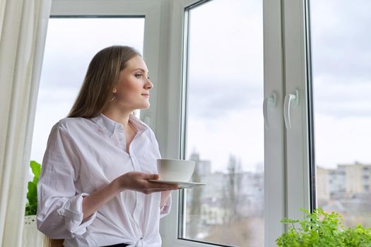 Beautiful smiling young woman with natural blond long hair holding cup coffee near panopam window. Attractive female in white shirt, winter spring season in window, copy space