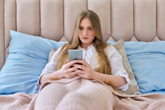 Serious young woman with smartphone resting lying at home in bed under a blanket reading surprised focused