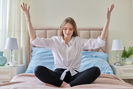 Yoga meditation at home, young beautiful blonde woman sitting in lotus position in bed, resting and meditating alone with closed eyes, raised her hands up