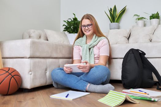 Portrait of teenage girl 12, 13 years old sitting at home on floor with school books, digital tablet. Online studies, e-education, resting schoolgirl using Internet modern digital technologies gadgets