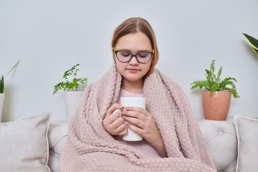 Cold autumn winter season at home, teenage girl under warm knitted blanket sitting on sofa with mug of hot tea