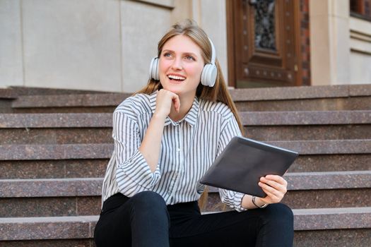 Young beautiful woman in headphones with digital tablet resting sitting on steps in city, listening music watching video. Happiness, relaxation, lifestyle, youth and technology