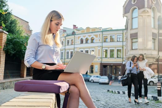 Beautiful young woman sitting outdoors using laptop, city street background. Female in business clothes, study, business, lifestyle concept