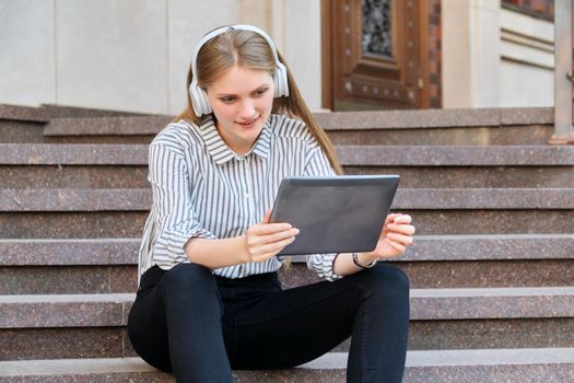 Young beautiful woman in headphones with digital tablet resting sitting on steps in city, listening music watching video. Happiness, relaxation, lifestyle, youth and technology