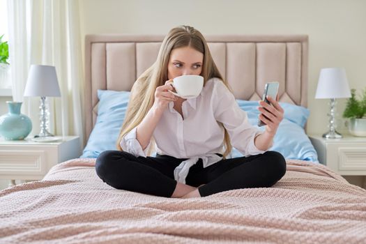 Happy talking young woman using video communication on smartphone, sitting at home in bed with cup of tea, relaxing. Rest, leisure, lifestyle, technology, telecommunications, people concept