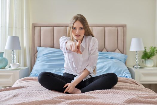 Confident smiling young beautiful woman showing index finger with hand gesture at you, female sitting at home in bed in bedroom