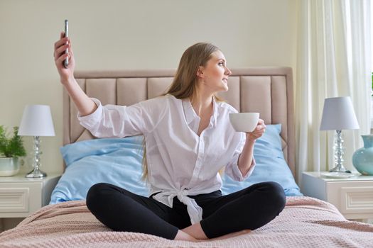 Happy talking young woman using video communication on smartphone, sitting at home in bed with cup of tea, relaxing. Rest, leisure, lifestyle, technology, telecommunications, people concept