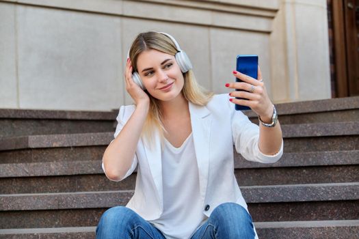 Young beautiful woman in headphones with smartphone resting sitting on steps in city, listening music watching video. Happiness, relaxation, lifestyle, youth and technology