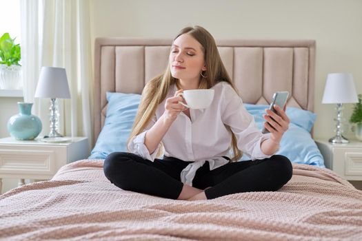 Happy talking young woman using video communication on smartphone, sitting at home in bed with cup of tea, relaxing. Rest, leisure, lifestyle, technology, telecommunications, people concept