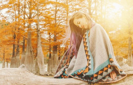 Boho style young woman wearing in poncho resting on tree trunk near the cypresses in autumn on sunny day.