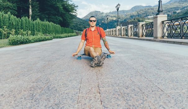 Fashionable tattoed guy in sunglasses sitting on longboard outdoor.