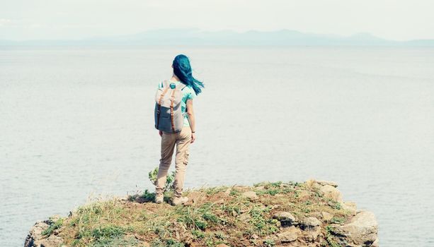 Backpacker young woman standing on peak of mountain and enjoying view of sea in summer, rear view.