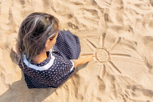 Girl draws a sun with sunbeams on the sand with her finger, top view.