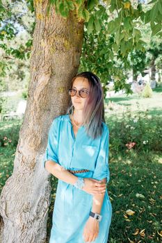 Beautiful young woman wearing in blue dress and sunglasses standing near a tree in summer park.