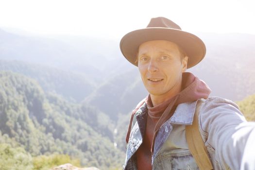 Smiling young man in a hat taking selfie on background of mountain valley, point of view.